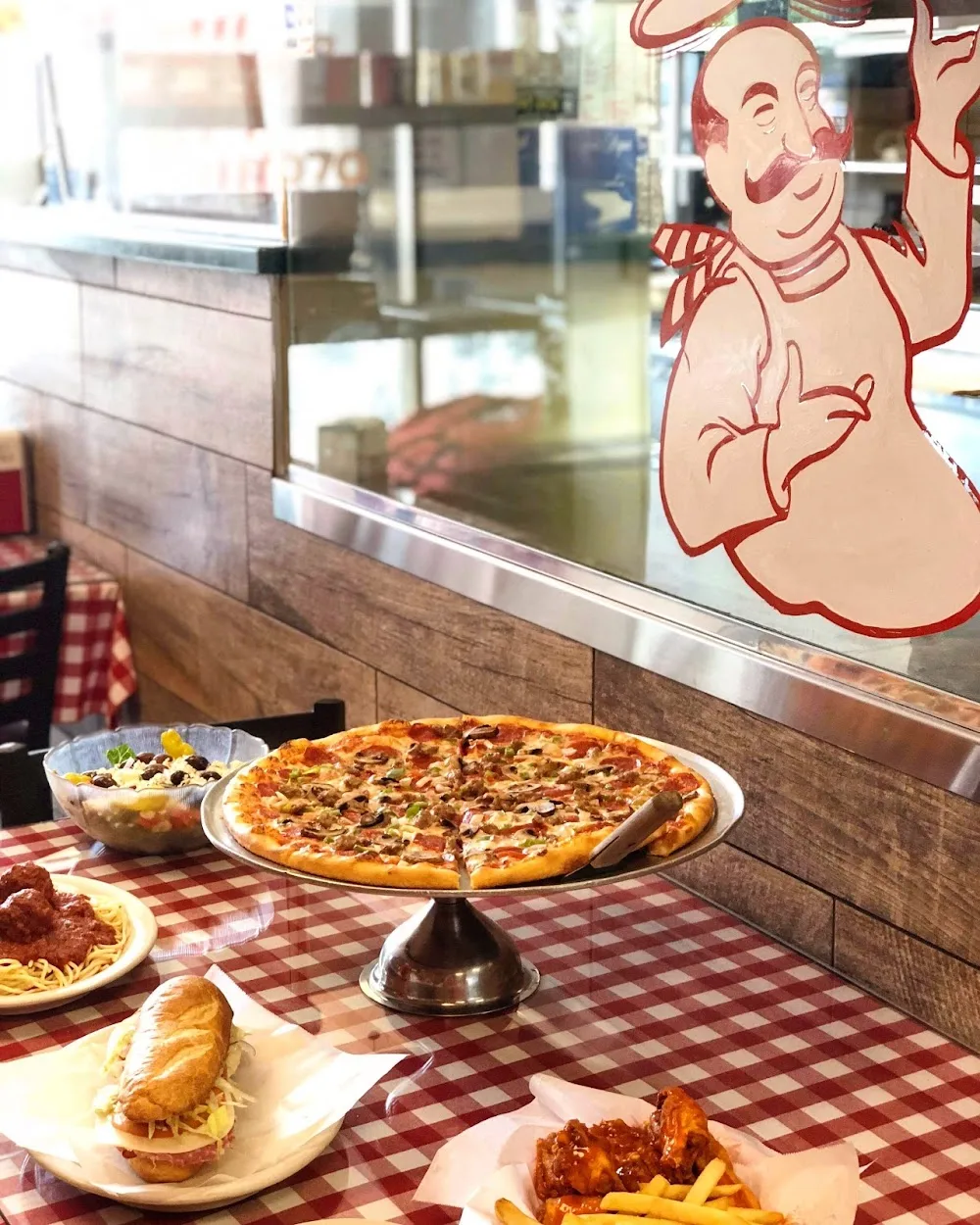 A table at Anthony's set with a whole pizza on a stand, a salad, spaghetti and meatballs, a sub and wings with fries, on a red checkered tablecloth